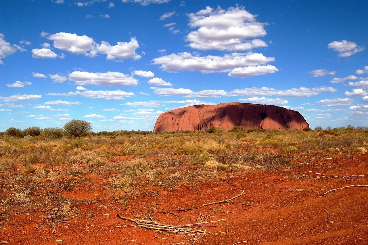 Uluru en el horizonte sobre tierra roja y arbustos dispersos